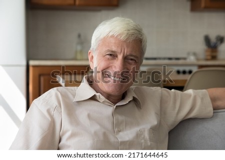 Handsome happy older grey-haired single man relaxing on cozy sofa smiling looking at camera enjoy carefree retired life, spend leisure alone at modern home. Untroubled male pensioner portrait concept