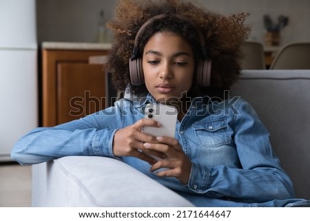 Beautiful African teenager girl wear wireless headphones using modern smartphone sit on sofa at home, watching vlog, videos on internet spend free time on-line. Young gen gadgets overuse, tech concept Royalty-Free Stock Photo #2171644647