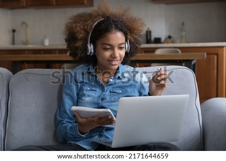 Attentive African teen-age girl studying at home, listens audio through wireless headphones, make task use laptop, engaged in easy and effective distancing on-line class. Video call, e-learn concept Royalty-Free Stock Photo #2171644659