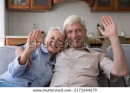 Middle-aged couple sit on sofa look at cam wave hands greeting friend, relatives or children living abroad start videocall talk use modern tech, enjoy virtual meeting at home. Video call event concept Royalty-Free Stock Photo #2171644679