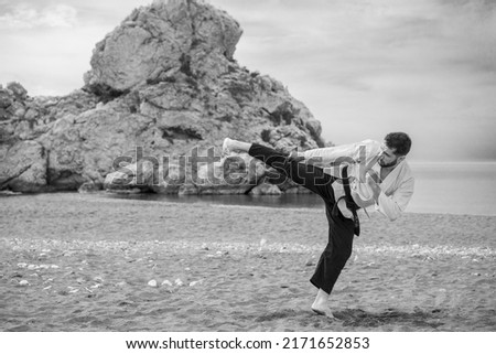 black and white image of a kung fu master in kimono wearing a black belt with the word "Bushido" written in Japanese performing a side kick on the sandy beach with a large rock in the background. Royalty-Free Stock Photo #2171652853