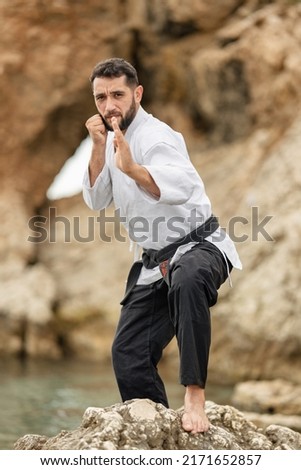 Martial arts master in kimono wearing a black belt with the word "bushido" in Japanese on a rock in a defensive stance. Royalty-Free Stock Photo #2171652857