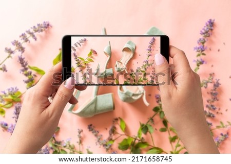 Woman taking photo of pastel mint green sandals shoes and meadow flowers with smartphone. Blogger, influencer or stylist capturing fashion footwear for social media. Pink background.