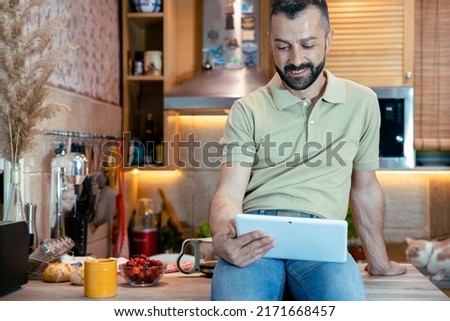 Bearded Man sitting on kitchen counter and looking at tablet computer. Male freelancer checking news in the morning at home