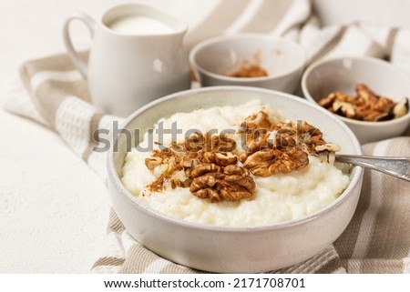 Creamy rice pudding whith milk, cinnamon powder and walnuts in white bowl with spoon on white table. Rice porridge with milk and topping for breakfast Royalty-Free Stock Photo #2171708701