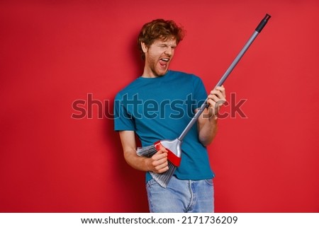 Playful young redhead man using broom like guitar while against red background