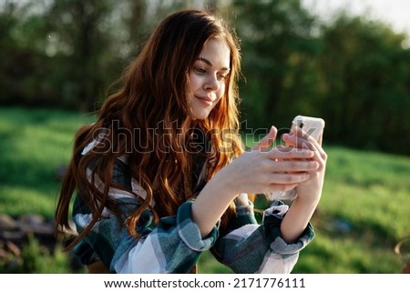A beautiful woman relaxing and working on her phone sitting in nature in the park among the trees smiling and holding her smartphone in her hand lit by the bright sunset light Royalty-Free Stock Photo #2171776111