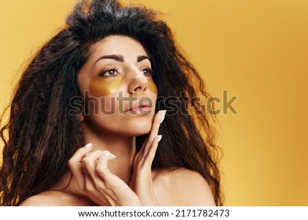 Curly pretty Latin woman in gold hydrogel patches under eye touch flawless healthy skin look aside posing isolated over yellow background. Anti-ageing skincare procedure concept Cosmetic Copy space