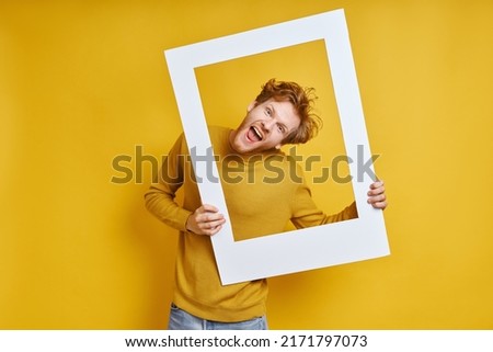 Playful redhead man looking through a picture frame while standing against yellow background