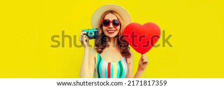 Summer portrait of happy smiling young woman with film camera and red heart shaped balloon wearing straw hat on yellow background, blank copy space for advertising text