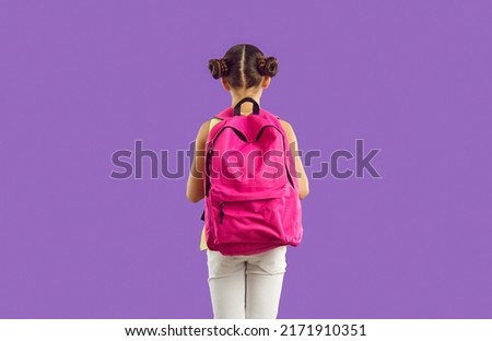 Back to school. New roomy pink school backpack on back of little girl, isolated on purple background. Schoolgirl with hairstyle with two bundles demonstrates new collection of school bags. Advertising Royalty-Free Stock Photo #2171910351