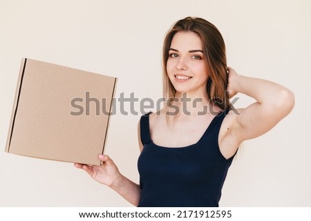 Young Girl in Black Dress Holds Cardboard Box in Her Hand Sideways. Caucasian Woman advertises box. Warm photo on beige background