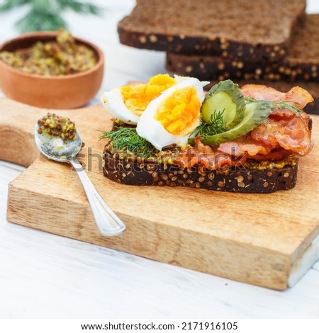 Delicious sandwich of rye bread with coriander with fried bacon, pickled cucumber, egg, dill and grainy mustard. A gourmet snack on a wooden board. Selective focus, square picture