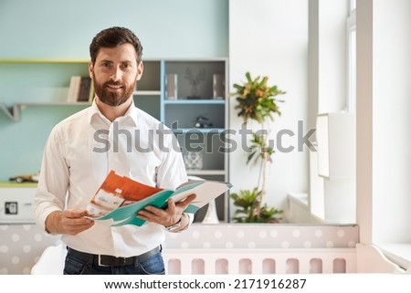 Smiling male sales manager holding catalogue of various furniture, while standing in light showroom. Portrait of bearded customer flipping brochure, while choosing goods. Concept of interior design. Royalty-Free Stock Photo #2171916287