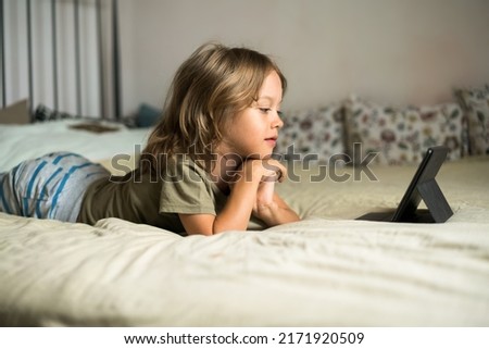 Boy lying on the bed using digital tablet computer playing games or watching cartoons at home. Child internet safety concept. Kid using electronic equipment. Parental control. Online education.