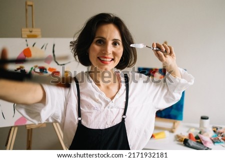 Real Female Artist Dirty with Paint, Wearing Apron, Crosses Arms while Holding Brushes, Looks at the Camera with a Smile. Authentic Creative Studio with Large Canvases. Head and Shoulders Portrait