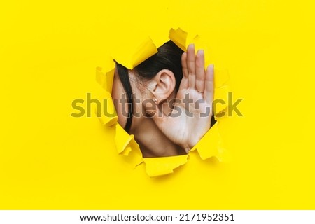 Close-up of a left woman's ear and hand through a torn hole in the paper. Bright yellow background, copy space. The concept of eavesdropping, espionage, gossip and tabloids. Royalty-Free Stock Photo #2171952351