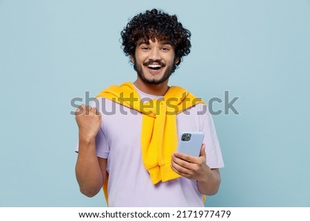 Happy young bearded Indian man 20s wears white t-shirt hold in hand use mobile cell phone doing winner gesture celebrate clenching fists isolated on plain pastel light blue background studio portrait Royalty-Free Stock Photo #2171977479