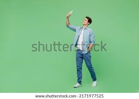 Full size body length side view young brunet man 20s years old wears blue shirt doing selfie shot on mobile cell phone post photo on social network isolated on plain green background studio portrait