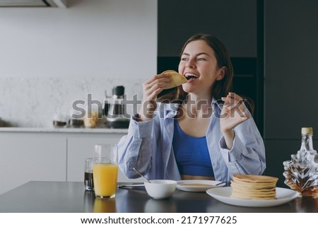 Young excited satisfied housewife woman 20s wearing casual clothes blue shirt eating breakfast pancakes with maple syrup cooking food in light kitchen at home alone. Healthy diet lifestyle concept. Royalty-Free Stock Photo #2171977625