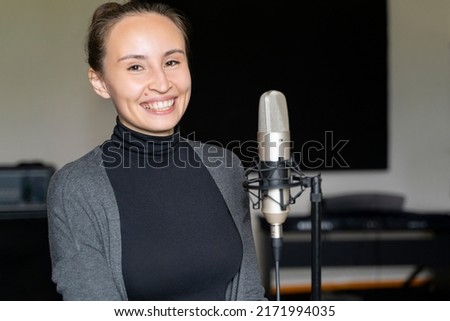 Portrait of young Chinese, Japanese or Caucasian brunette woman in casual clothes standing near retro vintage mic, smiling in music or broadcasting radio studio. Recording voice, talking to listeners