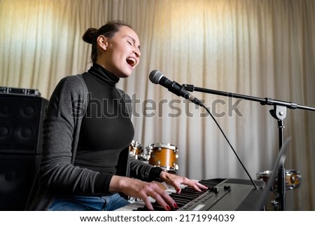 Young Asian or Caucasian brunette woman having fun singing song on mic emotionally loud with pleasure and playing digital keyboard electronic piano in music studio or school, event or concert stage Royalty-Free Stock Photo #2171994039