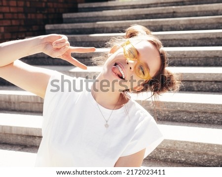 Young beautiful smiling hipster woman in trendy summer yellow jeans clothes. Carefree teen model posing in the street at sunset. Positive female outdoors. Cheerful and happy. Winks, shows peace sign