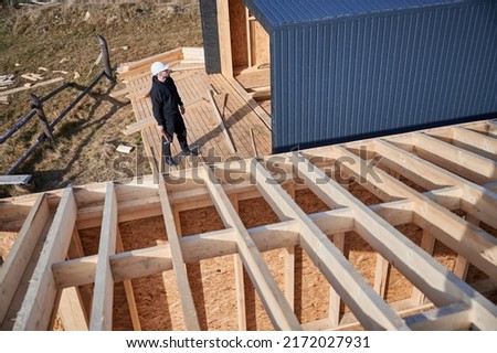 Male engineer building wooden frame house. Man developer on construction site, inspecting quality of work on sunny day, holding hammer in hands. Royalty-Free Stock Photo #2172027931