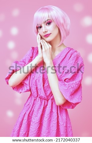 Beauty, fashion. Portrait of a cute teen girl with bright pink makeup and pink hair smiling dreamily and posing in fashionable pink dress. Pink background with lights. 