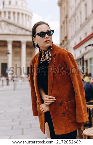 A young stylish girl with black hair in glasses and a stylish brown jacket walks around the city. Street walk, city life. Urban style girl. Lifestyle outdoor city portrait.