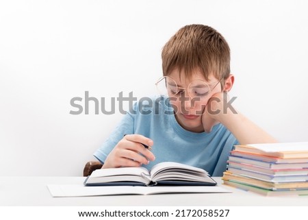 Bored boy doing homework at the desk. Schoolboy in glasses supports head with hand. Home schooling