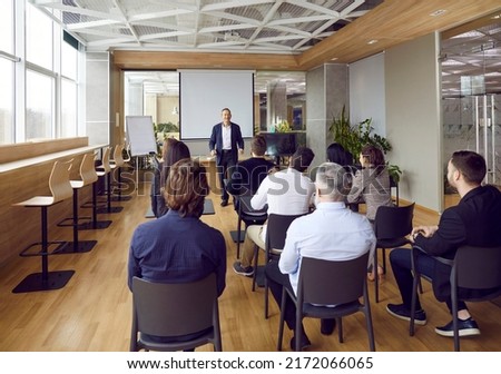 People working in joint office listen to male manager who talks about company's corporate program. Rear view of various businesspeople sitting at tables in office and listening to man near whiteboard. Royalty-Free Stock Photo #2172066065