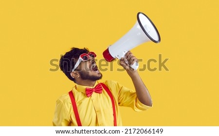 Screaming in megaphone. Funny Indian man in humorous clothes makes loud advertisement on vivid yellow background. Guy in yellow shirt, red suspenders and bow tie party glasses announces information