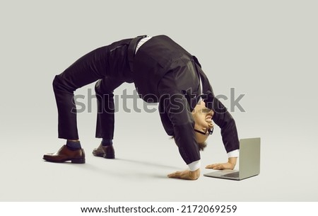 Happy energetic businessman practises yoga positions while working on laptop computer. Funny fit flexible young man in suit and glasses doing bridge pose on studio floor while using modern notebook PC Royalty-Free Stock Photo #2172069259