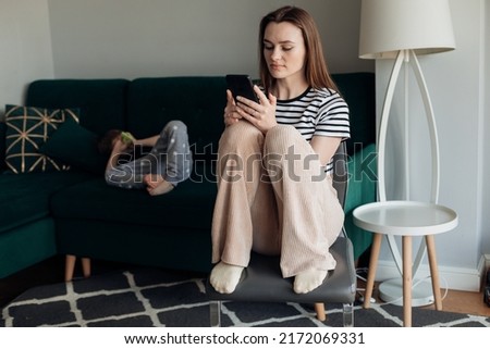Woman childminder sitting on chair and play telephone, little boy lying on big green sofa in living room. Indifference to child, idleness, bad work. Nanny not look after kid, bad relationships. Royalty-Free Stock Photo #2172069331