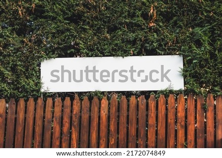 White blank billboard or advertising sign above a fence made of wood with pine trees behind