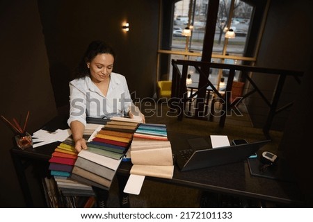 Charming dark-haired Hispanic woman, inspired interior designer selecting upholstery for new home design project, standing at a wooden desk with fabric samples and laptop in a cozy studio