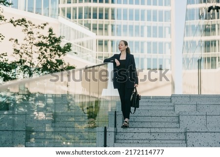 A female realtor is holding a bag while leaning on the glass parapet in a modern urban space on the stairs in the financial district. A businesswoman at sunset in a cluster of tall buildings. Royalty-Free Stock Photo #2172114777