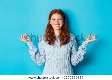 Smiling confident girl with red hair staying patient, holding hands in zen, meditation pose and staring at camera, practice yoga, standing calm against blue background