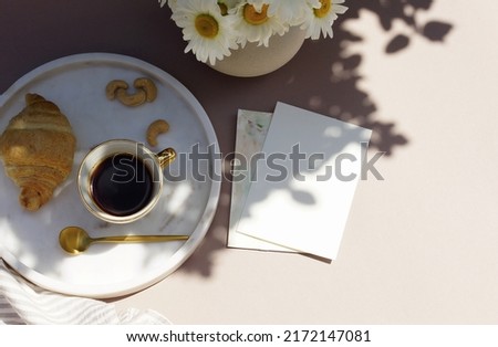 Blank paper sheet card mockup in  breakfast still life scene.Cup of coffee, croissant , daisy flowers in a vase top view on beige desk with sunlight shadows. Feminine lifestyle composition. 