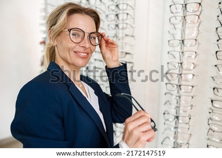 Woman chooses between two spectacle frames in an optician's shop and looks at the camera Royalty-Free Stock Photo #2172147519