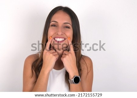 Happy young beautiful caucasian woman wearing white top over white background with toothy smile, keeps index fingers near mouth, fingers pointing and forcing cheerful smile