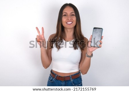 young beautiful caucasian woman wearing white top over white background holding modern device showing v-sign