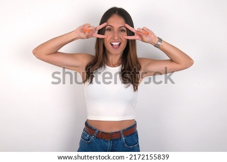 Cheerful positive young beautiful caucasian woman wearing white top over white background shows v-sign near eyes open mouth