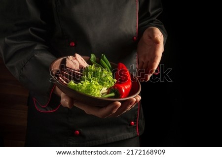 The chef is holding a plate with sliced steak and vegetables. Cooking concept on dark background with advertising space