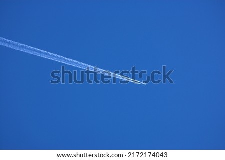 Airplane contrail against clear blue sky background with copyspace. View of a distant passenger jet plane flying on high altitude in blue sky leaving long white smoke trail behind. Air transportation Royalty-Free Stock Photo #2172174043