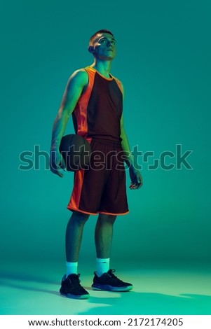 Full-length portrait of young man, professional basketball player in uniform with ball, posing isolated over blue background in neon light. Concept of sport, team game, action, active lifestyle, ad