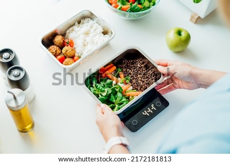 Healthy diet plan for weight loss, daily ready meal menu. Close up Woman weighing lunch box cooked in advance,ready to eat on kitchen scale. Balanced portion with healthy dish. Pre-cooking concept Royalty-Free Stock Photo #2172181813