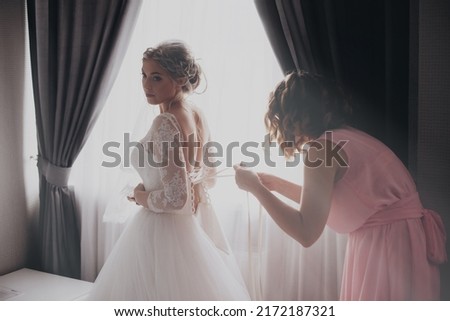 Bridesmaid helping slender bride lacing her wedding white dress, buttoning on delicate lace pattern with fluffy skirt on waist. Morning bridal preparation details newlyweds. Wedding day moments, wear. Royalty-Free Stock Photo #2172187321