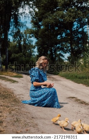 A woman in a long blue dress crouched next to ducklings in the woods.